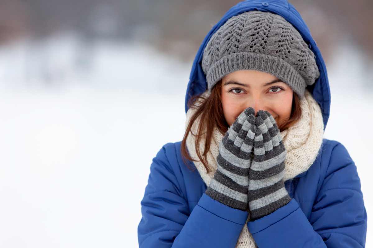 Donna in primo piano, vestita con un piumino blu con cappuccio, un cappello di lana grigio lavorato a maglia, una sciarpa color crema e guanti a righe grigie e bianche, che si copre il viso con le mani mentre è fuori sulla neve. Ha un'espressione sorridente e gli occhi che guardano in alto. Lo sfondo è un paesaggio invernale sfocato e nevoso.