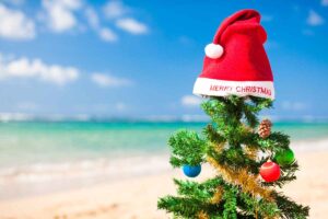 Primo piano di un piccolo albero di Natale decorato con palline colorate e un grande cappello di Babbo Natale con la scritta "Merry Christmas", posto su una spiaggia assolata con mare azzurro in lontananza. Natale al mare.