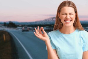 Una donna sorridente e allegra, vestita con una maglietta azzurra, sta tirando un filo di chewing gum fuori dalla sua bocca mentre guida. È su una strada asfaltata che si estende verso lo sfondo sfocato di colline innevate al tramonto o all'alba. La donna si trova sul lato destro del fotogramma e il filo di gomma è teso tra i suoi denti e la sua mano destra. Un'auto è visibile sulla carreggiata lontana.