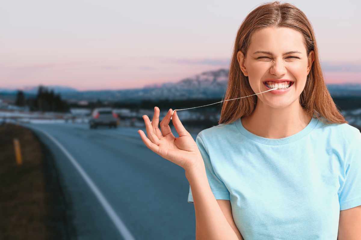 Una donna sorridente e allegra, vestita con una maglietta azzurra, sta tirando un filo di chewing gum fuori dalla sua bocca mentre guida. È su una strada asfaltata che si estende verso lo sfondo sfocato di colline innevate al tramonto o all'alba. La donna si trova sul lato destro del fotogramma e il filo di gomma è teso tra i suoi denti e la sua mano destra. Un'auto è visibile sulla carreggiata lontana.