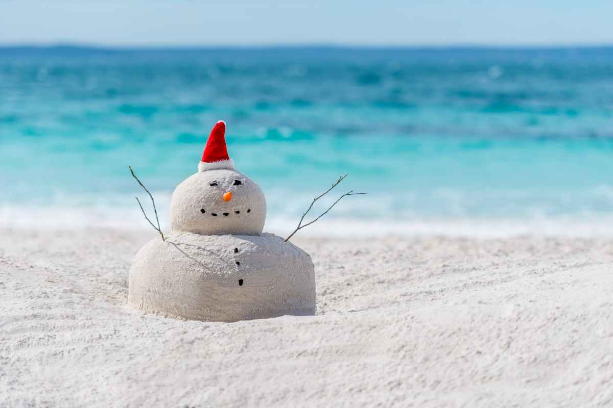Pupazzo di sabbia con cappello di Babbo Natale rosso su una spiaggia tropicale di sabbia bianca con acqua turchese sullo sfondo. 