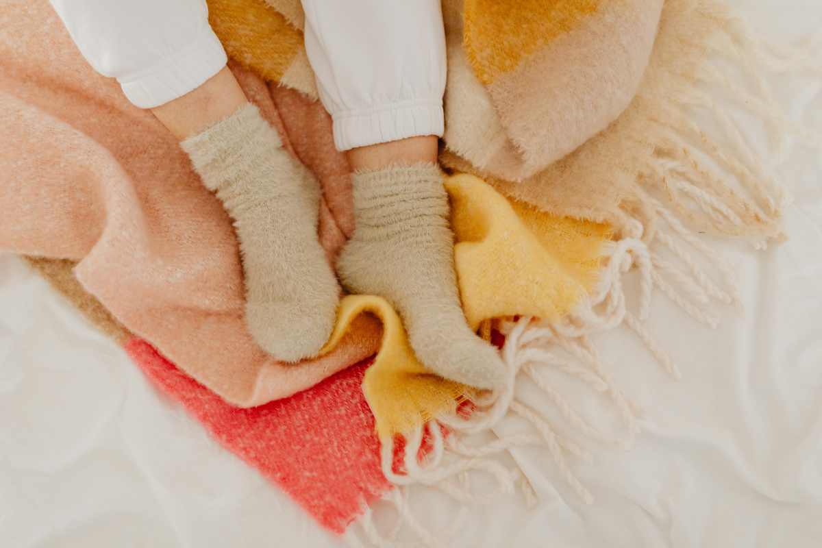 Primo piano dei piedi di una persona che indossa morbide calze di spugna color salvia chiaro, mentre è seduta su un letto con coperte e plaid pelosi di vari colori caldi: rosa, pesca, giallo e crema. La persona indossa pantaloni bianchi con elastico alla caviglia.