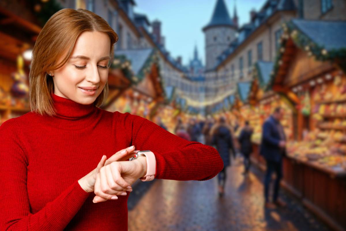 Una giovane donna bionda, con un maglione rosso a collo alto, guarda un orologio da polso rosa e argento che indossa. Lei è in primo piano, sorridente e con gli occhi chiusi, mentre lo sfondo è sfocato e mostra un mercatino di Natale affollato con bancarelle decorate, luci e persone che camminano. Si intravedono edifici tradizionali con tetti a punta.